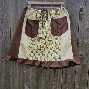 May include: A handmade skirt with a daisy floral pattern, brown pockets, and a ruffled hem. The skirt is hanging on a wooden hanger against a weathered wooden fence.