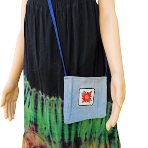 May include: A small, light blue denim crossbody bag with a blue strap. The bag features a square embroidered patch with a red flower design. The background includes a black and tie-dye dress.