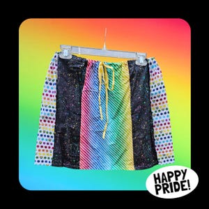 May include: A colorful skirt with a rainbow striped center panel, black side panels with paint splatters, and polka dot sleeves. The skirt has a yellow drawstring and is hanging on a hanger. The words "HAPPY PRIDE!" are in a white circle.