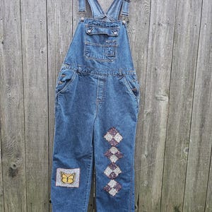 May include: Blue denim overalls with a butterfly patch and diamond-shaped patches on the legs. The overalls have adjustable straps, multiple pockets, and are hanging on a wooden hanger against a wooden fence.