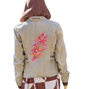 May include: A light brown jacket with a pink and floral lightning bolt patch on the back. The jacket has a brown hoodie attached to the collar.