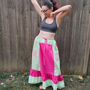 May include: A woman wearing a black sports bra and a long, multi-colored skirt with a pink and green patchwork design. The skirt has a ruffled hem and pockets on the sides.