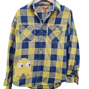 May include: Yellow and navy blue plaid button-down shirt with two chest pockets. A yellow Pac-Man character patch is sewn on the lower left side. The shirt is hanging on a wooden hanger.