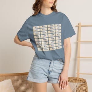 May include: A slate blue short-sleeve t-shirt featuring a repeating grid pattern of rectangular butter stick graphics. Each graphic is cream-coloured with blue text that reads BUTTER. The shirt has a crew neckline and a standard fit.