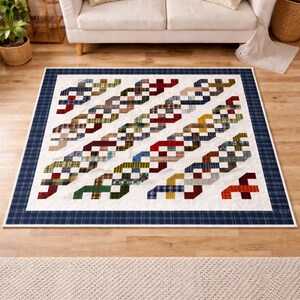 May include: A rectangular quilt featuring a white background with a repeating pattern of diagonal, interlocking ribbons made from various multicoloured plaid fabrics. The quilt is framed by a dark blue plaid border and has a visible quilted texture.