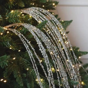 Peut inclure: Une branche décorative argentée recouverte de paillettes scintillantes, composée de multiples tiges fines et flexibles. Les longues branches en cascade sont disposées pour retomber vers le bas, créant un effet brillant sur les aiguilles vertes.