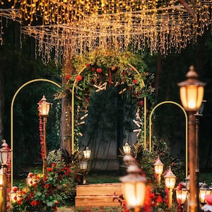 May include: Three gold metal arch frames arranged in a row, decorated with green foliage and red flowers. The central arch features hanging scrolls and is illuminated by warm string lights draped overhead, with vintage-style lanterns placed nearby.