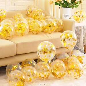 May include: A collection of clear latex balloons filled with metallic gold confetti. The balloons are scattered across a light-colored sofa and the floor, creating a festive display. Each balloon is transparent, showcasing the shiny gold circular confetti pieces inside, which vary in density and distribution throughout the interior of the balloons.
