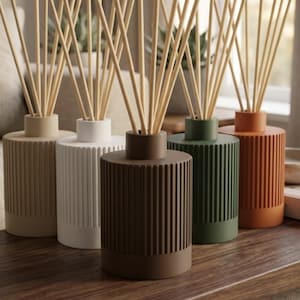 Op de afbeelding: Vijf cilindrische geurstokjes met verticale geribbelde texturen in beige, wit, donkerbruin, bosgroen en terracotta oranje. Elke fles heeft een smalle hals met een bundel lichtgekleurde houten stokjes die naar boven uitsteken.