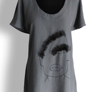 May include: A gray linen dress with a black embroidered face design. The dress has a scoop neck and short sleeves. The hemline is slightly curved.