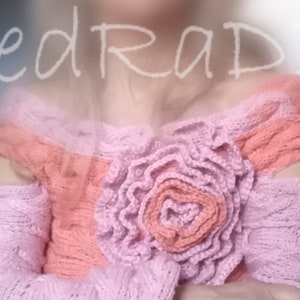 May include: A pink and peach knitted sweater with a large flower detail on the front. The sweater has a loose fit and off-the-shoulder style.