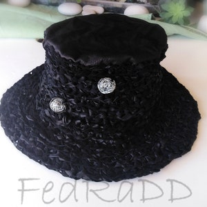 May include: Black hat with a wide brim and a textured, ruffled design. The top of the hat is smooth, and it features two decorative silver-colored ball accents. The hat is a stylish accessory.