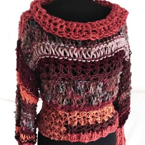 May include: A handmade, multi-colored crochet sweater displayed on a black mannequin. The sweater features a wide, open neckline and long sleeves. The color palette includes shades of red, brown, orange, and gray, creating a textured, unique design.