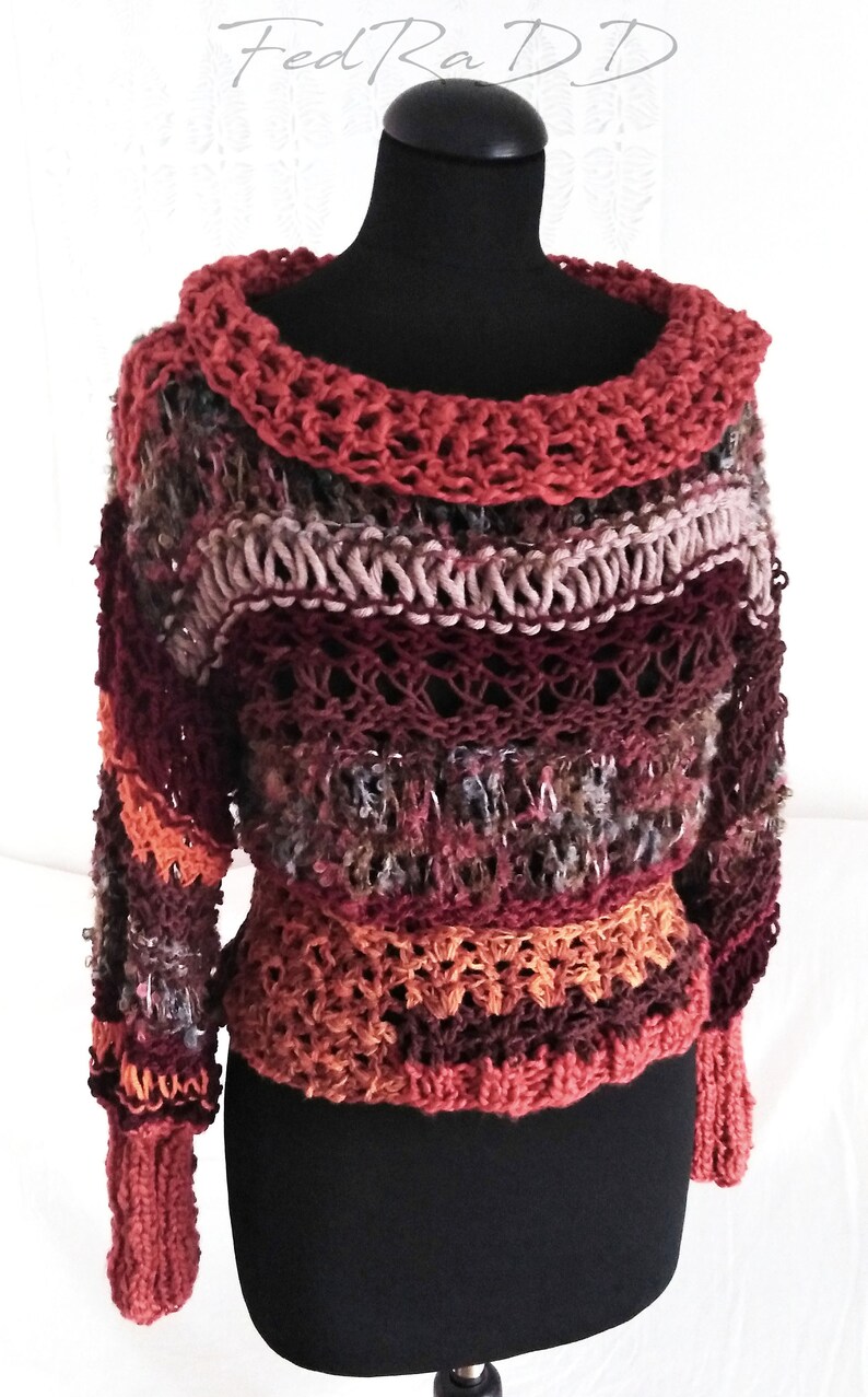 May include: A handmade, multi-colored crochet sweater displayed on a black mannequin. The sweater features a wide, open neckline and long sleeves. The color palette includes shades of red, brown, orange, and gray, creating a textured, unique design.