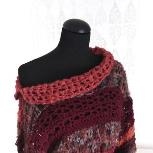 May include: A handmade, off-the-shoulder crochet poncho in shades of red, burgundy, and brown. The poncho features a wide, open-weave collar and a textured, multi-colored body. The garment is displayed on a black mannequin.