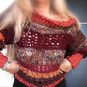 May include: A handmade, off-the-shoulder crochet sweater in shades of burgundy, orange, and brown. The sweater features a textured design with openwork sections and ribbed cuffs. The neckline and cuffs are a solid orange color.