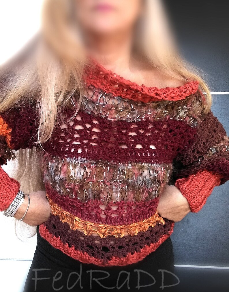 May include: A handmade, off-the-shoulder crochet sweater in shades of burgundy, orange, and brown. The sweater features a textured design with openwork sections and ribbed cuffs. The neckline and cuffs are a solid orange color.