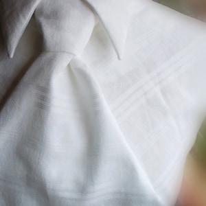 May include: A white linen shirt with a crisp collar and a tied neck. The shirt has a subtle texture and is made of high-quality fabric. The text "DDIVNA HANDMADE CLOTHES" is visible at the bottom of the image.