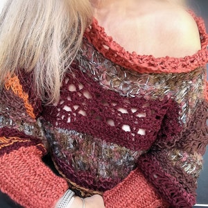 May include: A handmade, off-the-shoulder sweater in shades of burgundy, orange, and brown. The sweater features a crocheted neckline and sleeves, with a textured knit pattern. The sleeves are a solid coral color. The sweater is a unique, bohemian style.