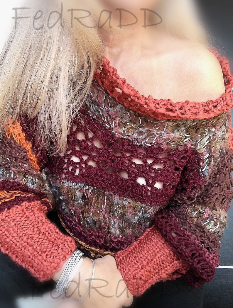 May include: A handmade, off-the-shoulder sweater in shades of burgundy, orange, and brown. The sweater features a crocheted neckline and sleeves, with a textured knit pattern. The sleeves are a solid coral color. The sweater is a unique, bohemian style.