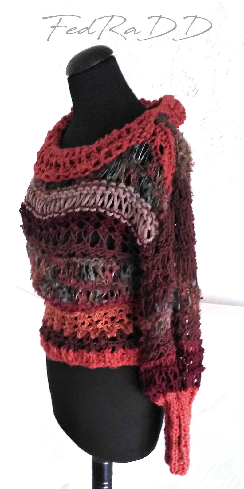 May include: A handmade, multi-colored crochet sweater displayed on a black mannequin. The sweater features a variety of colors, including burgundy, rust, and gray. The neckline and cuffs are a solid rust color. The text "Fed Ra D D" is in the upper left corner.