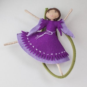 May include: A purple felt doll with a green felt hoop. The doll has a purple dress with a white belt and a white fringe around the bottom. The doll has brown hair and a painted face.