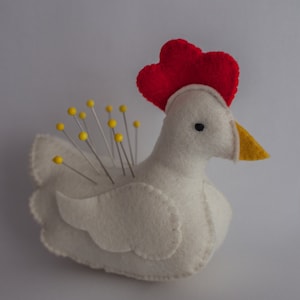 Puede incluir: Alfiletero de fieltro blanco en forma de pollo con una cresta de fieltro rojo y un pico de fieltro amarillo. El pollo tiene un ojo negro y está lleno de alfileres con cabezas amarillas.
