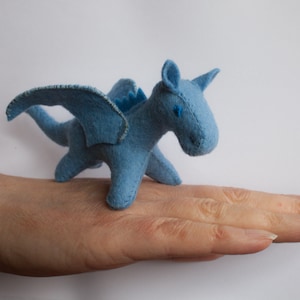 Könnte beinhalten: Ein kleiner blauer Filzdrache mit Flügeln, der auf einer Hand steht. Der Drache hat blaue Augen und ein aufgenähtes Lächeln.
