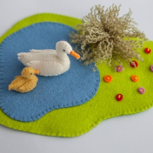 Peut inclure: Un ensemble de jeu en feutre avec un étang, un canard blanc, un caneton jaune et une zone herbeuse verte avec des fleurs colorées.