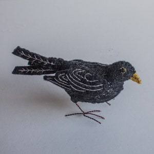 以下が含まれることがあります： 白と黒のニードルフェルトでできた鳥。黄色いクチバシとワイヤー製の足がついています。鳥は白い表面の上に立っています。