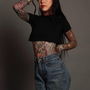 Può includere: Un top corto nero a maniche corte abbinato a jeans blu a vita alta. Il top presenta un semplice scollo rotondo e un orlo dritto che termina sopra la vita, mentre i jeans hanno una classica chiusura con bottone e cerniera lampo.