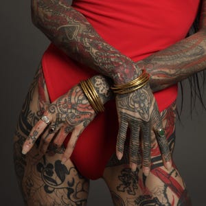 Op de afbeelding: Een felrode bodysuit gecombineerd met meerdere dunne goudkleurige metalen armbanden om beide polsen. Aan de vingers zijn drie verschillende ringen te zien, waaronder een met een donkere steen en een met een groene steen.