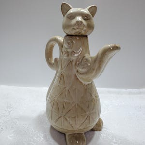 以下が含まれることがあります： 光沢のある仕上げの猫の形をしたベージュの陶器製ティーポット。本体には盛り上がった幾何学的なダイヤモンド模様と渦巻き模様が施されています。湾曲したハンドル、突き出た注ぎ口、猫の頭を形成する取り外し可能な蓋が付いています。