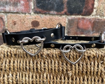 May include: Two black leather choker necklaces with silver heart-shaped hardware. The necklaces are on a woven wicker surface.