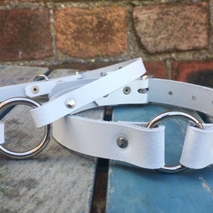 May include: Two white leather collars with silver metal rings. The collars are on a wooden surface.
