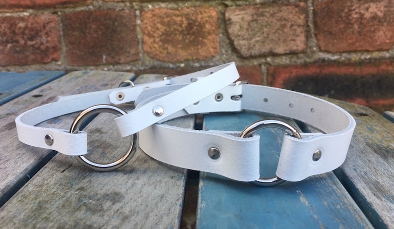 May include: Two white leather collars with silver metal rings. The collars are on a wooden surface.