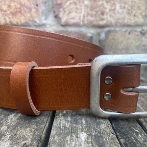 May include: A tan leather belt featuring a smooth finish, a single leather keeper loop, and a large, rectangular, brushed silver-tone metal buckle secured by two visible metal rivets on the strap near the buckle attachment point.