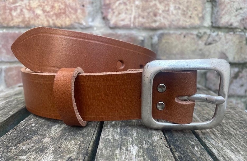 May include: A tan leather belt featuring a smooth finish, a single leather keeper loop, and a large, rectangular, brushed silver-tone metal buckle secured by two visible metal rivets on the strap near the buckle attachment point.