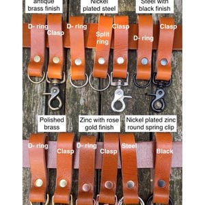 May include: A collection of tan buffalo leather belt loops featuring various metal hardware attachments including D-rings, clasps, split rings, and spring clips in finishes such as antique brass, nickel, black, polished brass, and rose gold.