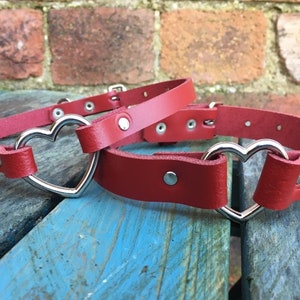 May include: Two red leather choker necklaces with silver heart-shaped clasps. The necklaces are on a wooden surface.