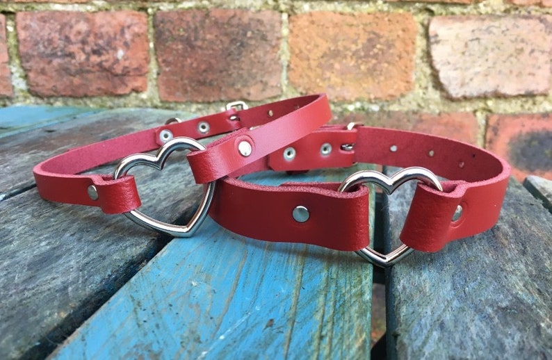 May include: Two red leather choker necklaces with silver heart-shaped clasps. The necklaces are on a wooden surface.