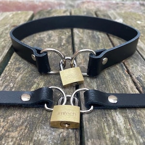 May include: Black leather collar and matching restraint with silver-tone metal rings and brass padlocks. The padlocks are engraved with the word "amtech". The items are displayed on a weathered wooden surface.
