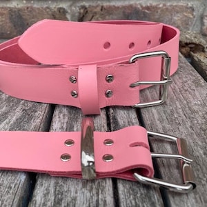 May include: Two pink leather belts with silver-colored buckles. The belts are displayed on a weathered wooden surface. One belt is coiled, and the other is laid out flat, showcasing the buckle and the belt's holes.