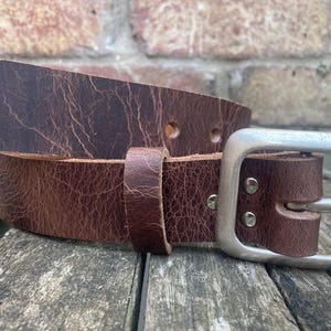 May include: A dark brown leather belt with a distressed, cracked texture. The belt features a rectangular, brushed silver-tone metal buckle secured by two rivets and a matching leather keeper loop to hold the strap in place.