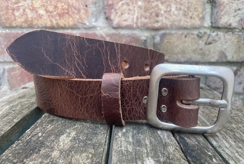 May include: A dark brown leather belt with a distressed, cracked texture. The belt features a rectangular, brushed silver-tone metal buckle secured by two rivets and a matching leather keeper loop to hold the strap in place.