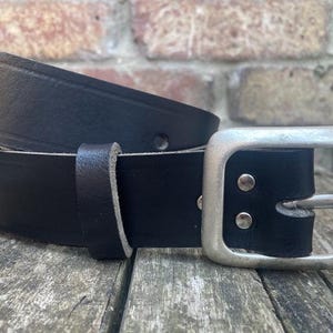 May include: A black leather belt featuring a smooth finish and a rectangular silver-toned metal buckle. The belt includes a matching leather keeper loop and is secured with two silver-toned metal rivets near the buckle attachment point.