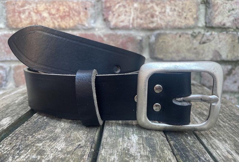 May include: A black leather belt featuring a smooth finish and a rectangular silver-toned metal buckle. The belt includes a matching leather keeper loop and is secured with two silver-toned metal rivets near the buckle attachment point.