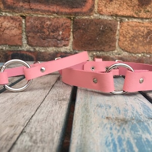 May include: Two pink leather collars with silver metal rings. The collars are on a wooden surface.