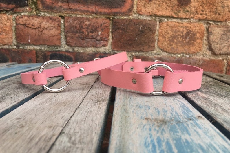May include: Two pink leather collars with silver metal rings. The collars are on a wooden surface.