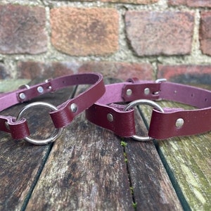 May include: Two burgundy leather collars with silver metal rings. The collars are lying on a wooden surface.